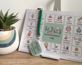Puede incluir: Un tapete de comunicación con varias ilustraciones de necesidades y sentimientos, como "Tengo hambre" y "Quiero un abrazo". Un pequeño juego de "Mis tarjetas de comunicación" está adjunto. A la izquierda hay una planta suculenta en maceta.