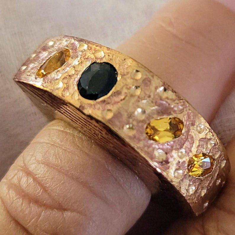 Puede incluir: Un anillo de oro con una superficie texturizada y tres piedras preciosas. Las piedras preciosas son un zafiro negro y dos citrinos amarillos.