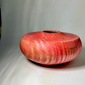 Op de afbeelding: Een handgemaakte houten kom in levendige roze en rode tinten. De kom heeft een afgeronde vorm met een brede, platte basis en een cirkelvormige opening. De houtnerf is zichtbaar en voegt textuur toe. De kom heeft een diameter van ongeveer 15 cm.