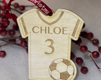 May include: A wooden Christmas ornament shaped like a soccer jersey with the name "CHLOE" and the number "3" on it. A soccer ball is on the bottom of the ornament.