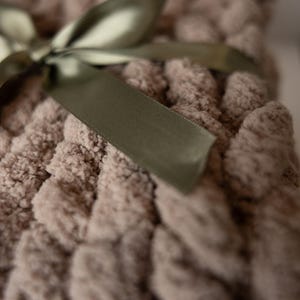May include: Close-up of a chunky knit blanket in a soft taupe color. A sage green satin ribbon is tied in a bow on top of the blanket. The blanket has a textured, looped design.