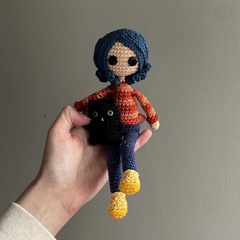 May include: Crocheted doll with blue hair, a striped orange and red sweater, blue pants, and yellow shoes. The doll is holding a black cat. The doll's eyes are black, and the skin is beige.