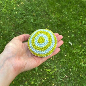 May include: A round, crocheted object featuring a spiral pattern of alternating light blue and lime green yarn. The item has a dense, textured stitch construction and a circular shape, displaying a consistent color rotation from the center outward.