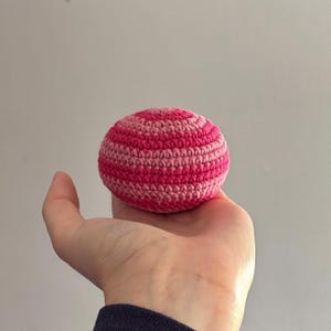 May include: A handmade, striped crochet ball in shades of pink and rose. The ball is held in a hand, showcasing the texture and color variations of the yarn. This item is a craft or toy.