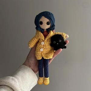 May include: A handmade crochet doll with blue hair, a yellow coat, and blue pants. The doll is holding a black creature. The doll has button eyes and a red shirt. The doll's shoes are yellow.