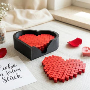Könnte beinhalten: Ein schwarzes herzförmiges Tablett und ein separates rotes pixeliges Herz werden auf einer hellen Oberfläche präsentiert. Das Tablett ist mit roten Blöcken gefüllt, und eine kleine Vase mit weißen Blumen und herzförmigen Kerzen befindet sich in der Nähe. Eine Karte mit Text ist ebenfalls vorhanden.