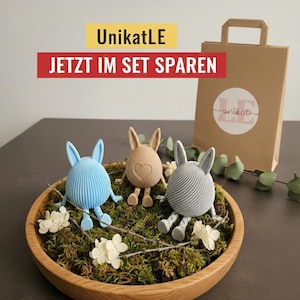 Könnte beinhalten: Drei dekorative Hasenfiguren in Blau, Beige und Grau, jede mit einem einzigartigen Design, sitzen in einer Holzschale mit Moos und weißen Blumen. Eine braune Papiertüte mit Logo befindet sich im Hintergrund. Der Text "UnikatLE" und "JETZT IM SET SPAREN" sind sichtbar.