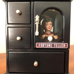 May include: Black wooden jewellery box with a fortune teller doll inside a glass window. The box has three drawers and a sign that says "Fortune Teller".