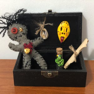 May include: A black wooden box containing a grey rag doll, a yellow face mask, a seashell, a small glass vial with green beads and a rolled up piece of parchment paper.