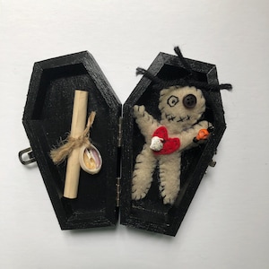 May include: A small black coffin with a hinged lid is open to reveal a voodoo doll with a red heart and a rolled up scroll. The doll is made of fabric and has a button for an eye.