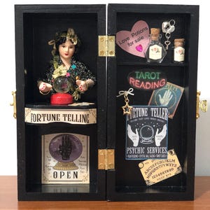 May include: A black wooden box with a fortune teller theme. The box is open and reveals a miniature fortune teller doll, a crystal ball, tarot cards, and other fortune telling items. The box has a sign that says "Fortune Telling" and another sign that says "Tarot Reading".