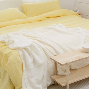 May include: A white linen throw blanket draped over a yellow linen bedspread. The bed is made with two yellow linen pillows. A wooden side table with two shelves is next to the bed.