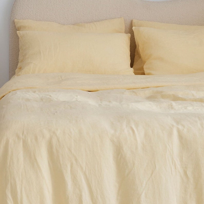 K&ouml;nnte beinhalten: Ein gelber Leinen-Bettbezug und zwei passende Kissenbez&uuml;ge. Die Bettw&auml;sche ist auf einem Bett mit einem hellbraunen Kopfteil drapiert.