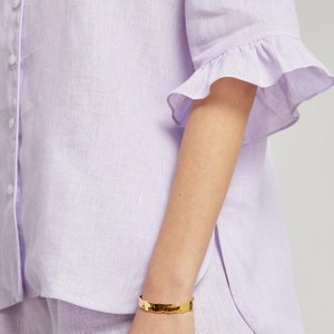 May include: A lavender linen shirt with a ruffled short sleeve and a button-up front.
