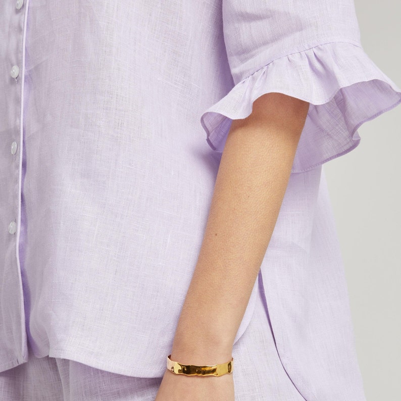 May include: A lavender linen shirt with a ruffled short sleeve and a button-up front.