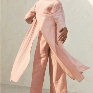 May include: A light pink linen pantsuit with a long, flowing jacket and wide-leg pants. The jacket has a shawl collar and a tie at the waist.