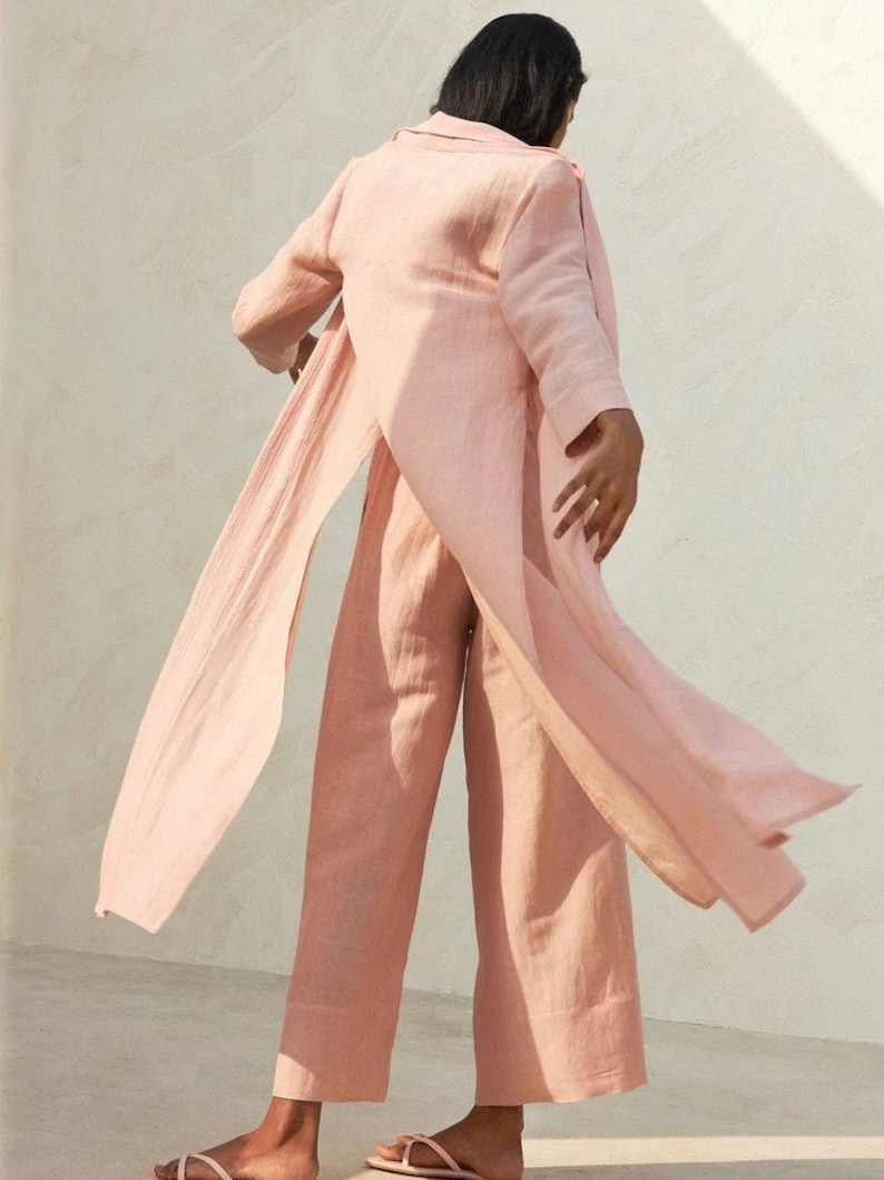 May include: A light pink linen pantsuit with a long, flowing jacket and wide-leg pants. The jacket has a shawl collar and a tie at the waist.