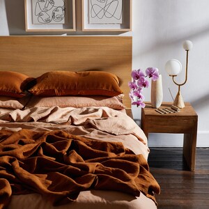 K&ouml;nnte beinhalten: Ein Bett mit einer braunen Bettw&auml;sche aus Leinen und einem Laken aus pfirsichfarbenem Leinen. Zwei braune Kissen aus Leinen liegen auf dem Bett. Ein Nachttisch aus Holz mit einer Vase mit rosa Orchideen und einer goldenen Lampe steht neben dem Bett.