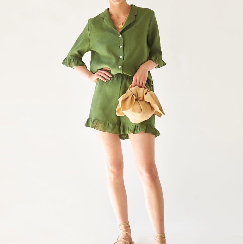 May include: A green linen shirt and matching shorts set with a ruffled hem. The shirt has a collar and button closure. The model is holding a small, light brown, fabric bag.