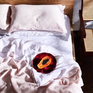 K&ouml;nnte beinhalten: Ein wei&szlig;es Leinen-Bettlaken mit einem rosafarbenen Leinen-Bettbezug. Ein roter Teller mit einer Papaya-Scheibe darauf liegt auf dem Bett.