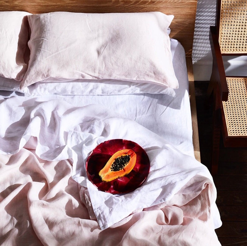K&ouml;nnte beinhalten: Ein wei&szlig;es Leinen-Bettlaken mit einem rosafarbenen Leinen-Bettbezug. Ein roter Teller mit einer Papaya-Scheibe darauf liegt auf dem Bett.
