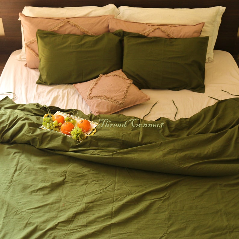 K&ouml;nnte beinhalten: Ein gr&uuml;ner Bettbezug mit einem wei&szlig;en Laken und mehreren Kissen. Die Kissen sind mit einem rosa Stoff mit Fransenbesatz bezogen. Ein silbernes Tablett mit Orangen und Trauben steht auf dem Bett.