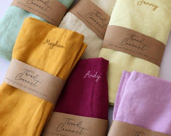 May include: A collection of six folded fabric napkins in light green, beige, yellow, mustard, burgundy, and lavender. Each napkin features an embroidered name and a brown paper band with the text Thread Connect POP IN THE RANGE OF CONSCIOUS LIVING.