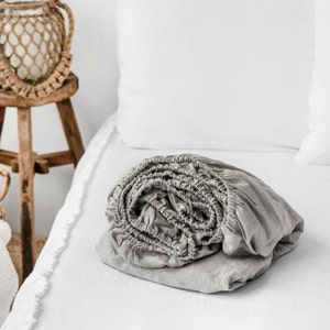 May include: A gray fitted sheet is rolled up and placed on a white bed. The sheet is made of a soft, linen fabric.