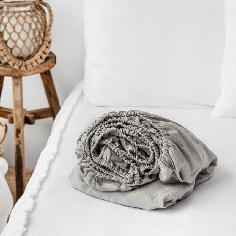 May include: A gray fitted sheet is rolled up and placed on a white bed. The sheet is made of a soft, linen fabric.