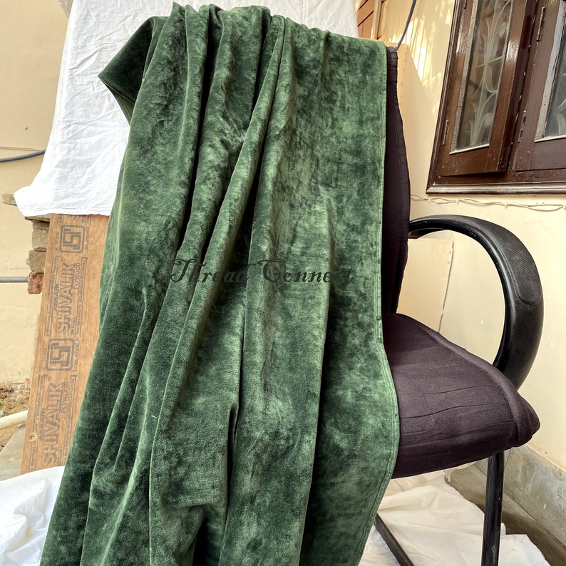 Pu&ograve; includere: Un tessuto di velluto verde foresta drappeggiato su una sedia nera. Il tessuto ha un aspetto morbido e strutturato con pieghe e ombre visibili. Il testo "Thread Connection" &egrave; visibile sul tessuto. Lo sfondo include una finestra e una tavola di legno.