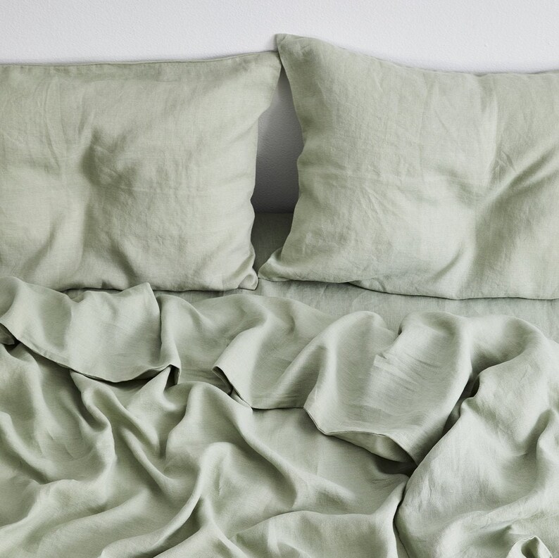 May include: A sage green linen bedding set with a flat sheet, two pillowcases, and a duvet cover. The bedding is wrinkled and has a relaxed, casual look.