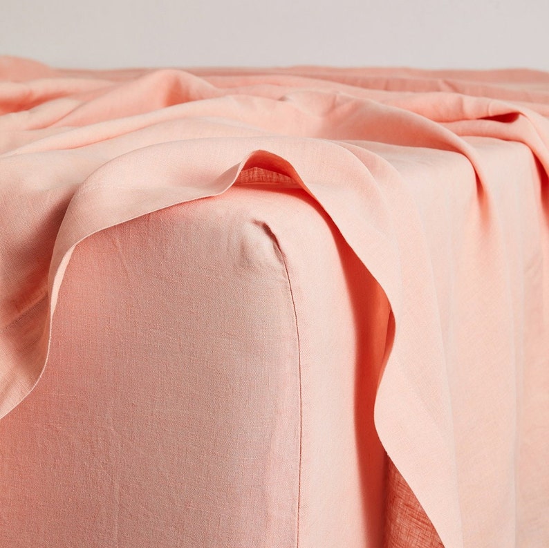May include: A peach-colored fitted sheet made of linen. The sheet is folded and draped over a corner of a mattress.