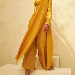 May include: A mustard yellow linen pantsuit with a long, draped jacket and a cropped top. The pants have a wide leg and the jacket is open in the front. The model is wearing the suit with a pair of beige sandals.