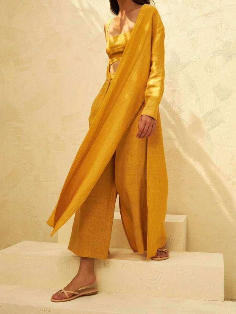 May include: A mustard yellow linen pantsuit with a long, draped jacket and a cropped top. The pants have a wide leg and the jacket is open in the front. The model is wearing the suit with a pair of beige sandals.