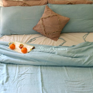 K&ouml;nnte beinhalten: Ein hellblaues Bettw&auml;sche-Set mit einem braunen Kissen mit Fransen. Die Bettw&auml;sche liegt auf einem Bett mit einer wei&szlig;en Matratze und einem Tablett mit Obst auf dem Bett.