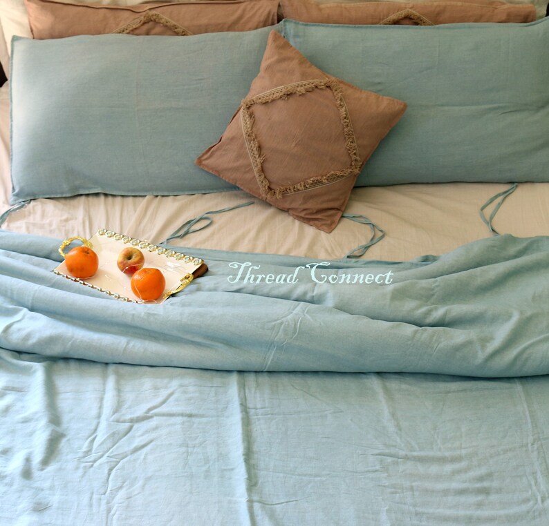 K&ouml;nnte beinhalten: Ein hellblaues Bettw&auml;sche-Set mit einem braunen Kissen mit Fransen. Die Bettw&auml;sche liegt auf einem Bett mit einer wei&szlig;en Matratze und einem Tablett mit Obst auf dem Bett.