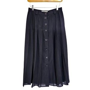 Peut inclure: Jupe midi noire avec devant boutonné et design plissé. La jupe est suspendue à un cintre en bois. La jupe a une taille haute et un style classique et intemporel.