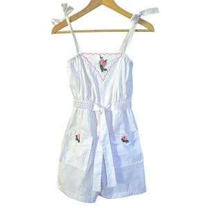 May include: White romper with tie straps and a matching belt. The romper features two front pockets and is adorned with embroidered pink roses. A pink and white embroidered floral design is on the bodice. The romper is hanging on a wooden hanger.