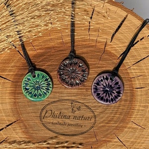 Può includere: Tre ciondoli in ceramica con cordini neri, ognuno con un colore e un design diverso. I ciondoli sono esposti su una superficie di legno con una spiga di grano sullo sfondo. Il testo "Philina naturi - handmade jewellery" è visibile sotto i ciondoli.