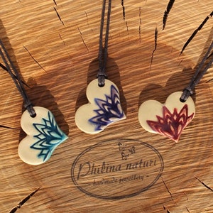 Könnte beinhalten: Drei Keramik-Herzanhänger mit filigranen Blumenmustern in Blautönen, Lila und Rot. Jeder Anhänger ist an einer braunen Schnurkette befestigt. Die Anhänger werden auf einer Holzoberfläche mit dem Text "Philina naturi - handmade jewellery" präsentiert.