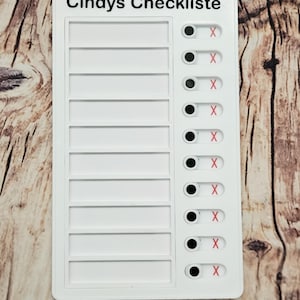 Könnte beinhalten: Eine weiße Checkliste mit dem Text "Cindys Checkliste" oben. Die Checkliste hat mehrere leere Kästchen für Einträge und eine Reihe von Kippschaltern mit roten "X"-Markierungen. Der Hintergrund ist eine Holzoberfläche.