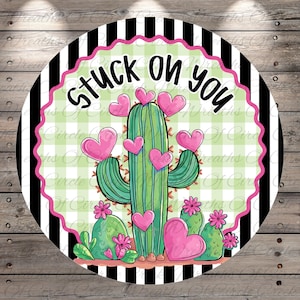 May include: Round sign with a green cactus decorated with pink hearts and flowers. The sign has the text "Stuck on you" in black script. The background features a black and white striped border and a green and white checkered pattern.