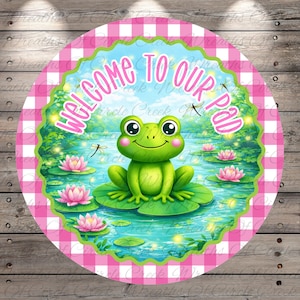 May include: A round sign with a pink and white checkered border. The center features a green frog sitting on a lily pad in a pond with pink water lilies. The text "WELCOME TO OUR PAD" is in pink script.