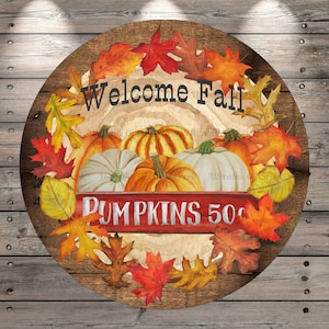 May include: A round wooden sign with a fall theme. It features a wreath of autumn leaves in shades of red, orange, and yellow surrounding a group of pumpkins. The sign also has the text "Welcome Fall" and "Pumpkins 50c" in white lettering.