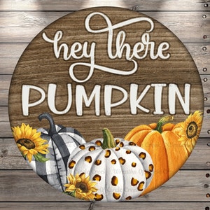May include: Round wooden sign with the text "Hey there Pumpkin" in white lettering. The sign is decorated with three pumpkins, one with black and white checkered pattern, one with white and brown leopard print, and one orange pumpkin. The pumpkins are surrounded by sunflowers.