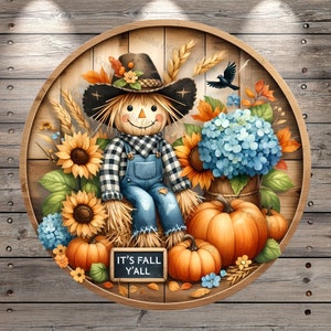 May include: A circular wooden sign featuring a scarecrow with a black and white checkered shirt and blue overalls, surrounded by sunflowers, pumpkins, and blue hydrangeas. The sign reads "IT'S FALL Y'ALL".