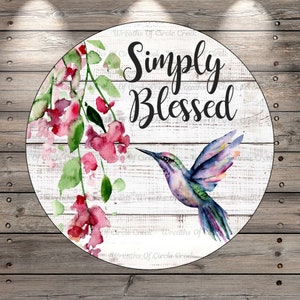 May include: A round wooden sign with a whitewashed background and a watercolor hummingbird and pink flowers. The sign says "Simply Blessed" in black script.