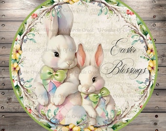 Könnte beinhalten: Ein rundes, grün umrandetes Schild mit einer Aquarell-Illustration von zwei weißen Kaninchen, die pastellfarbene Ostereier halten. Die Kaninchen sind von einem Kranz aus Blumen und Blättern umgeben. Der Text "Easter Blessings" ist in einer dekorativen Schrift geschrieben.