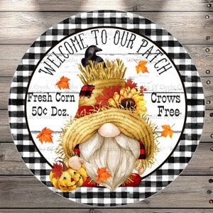 May include: A round, black and white checkered sign with the text "Welcome to Our Patch" in a white banner. The sign features a gnome wearing a straw hat with a scarecrow face, holding a jack-o-lantern. The text "Fresh Corn 50¢ Doz." and "Crows Free" is also on the sign.