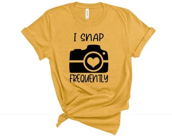 May include: Mustard yellow t-shirt with a black camera graphic and the text "I SNAP FREQUENTLY". The camera graphic includes a heart in the center of the lens. The shirt is knotted at the bottom.